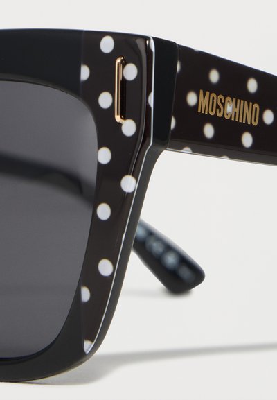 Branchage de lunettes de soleil noir avec des pois blancs et un logo Moschino doré, montrant une lentille teintée foncée et un détail de monture rectangulaire.