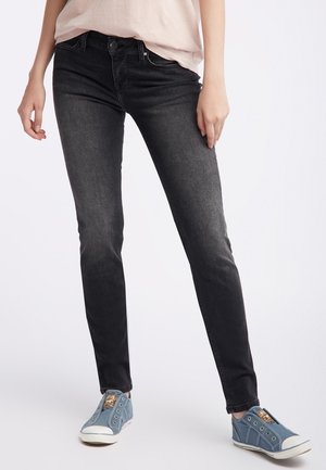 Femme portant un jean skinny noir délavé et des baskets bleues à enfiler, debout jambes écartées sur un fond uni.