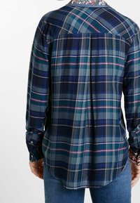 Persona con una camicia a quadri blu, verde e viola con ricami floreali sul colletto, abbinata a jeans blu, vista da dietro.