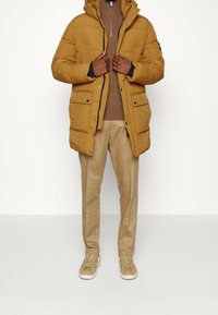Mosterdgele pufferjack met capuchon, voorzijde met rits, en meerdere zakken, gedragen over een bruine trui en beige broek, gecombineerd met beige sneakers.