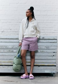 Creme zip-up hoodie, strukturerede lavendelshorts, pink platform klipklapper og en grøn polstret totebag. Sat mod en hvid murstensvæg.