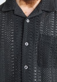 Svart hekletrøye med klippet krage, seks knapper foran og en enkelt brystlomme med en teksturert, åpenvevd design.