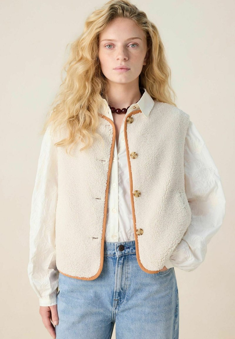 Gilet en polaire beige avec des bordures brunes, comportant quatre boutons, porté sur une chemise de couleur claire. Un jeans en denim bleu complète l'ensemble.