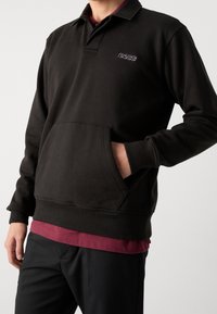 Černý pulovr s límečkem, s přední kapsou a žebrovaným lemem. Vyšívané logo "BOSS" ve fialové barvě na hrudi s vrstvenou vínově červenou košilí pod ním.