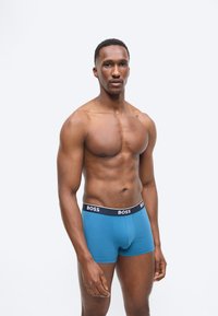 Αθλητικός άνδρας όρθιος, φοράει μπλε μποξεράκια BOSS με μαύρη ζώνη μέσης, μπροστά σε απλό ανοιχτό γκρι φόντο.