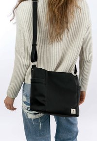 Schwarze Schultertasche mit gerippter Textur, verstellbarem Tragegurt und Fronttasche. Getragen mit einem weißen Strickpullover und zerrissenen blauen Jeans.