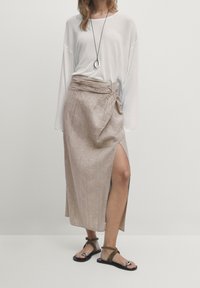 Jupe en lin beige légère avec un détail de taille nouée et une fente haute sur la cuisse ; associée à un haut blanc ample et des sandales plates.