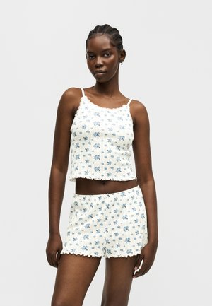 Kvinde iført hvid ribbet tanktop og shorts med små blå blomsterprint, stående mod en ensartet lys baggrund.