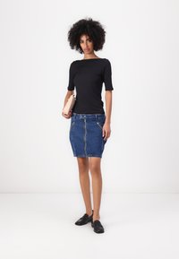 Svart tettsittende topp med korte ermer, kombinert med en blå denimskjørt med glidelås foran og to sidelommer med glidelås. Svart loafers og en beige clutch.