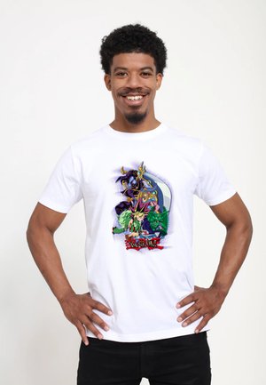 Uśmiechnięty mężczyzna z rękami na biodrach w białej koszulce z kolorowymi postaciami i logo Yu-Gi-Oh! na przodzie na jednolitym tle.