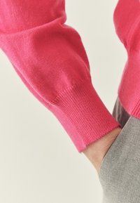 Pull rose vif avec poignets côtelés, présentant une texture douce et un design sans coutures. Une main repose sur une poche grise.