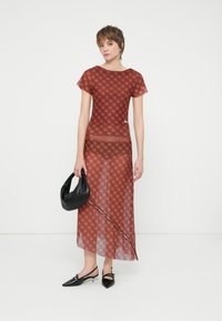 Red fitted dress with geometric pattern, sheer overlay, asymmetrical hem, short sleeves. Model holds a black handbag, paired with black shoes.