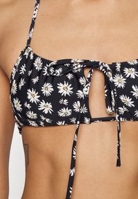 Top de bikini preto com padrão de margaridas brancas, tecido franzido, alças ajustáveis e detalhe de laço central. Apresenta um design com recorte em forma de olho-chave.