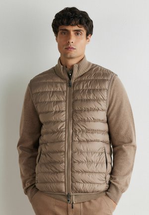 Mężczyzna w jasnobrązowej pikowanej kamizelce bez rękawów na beżowym swetrze, stojący oparty o gładką białą ścianę, z rękami w kieszeniach.