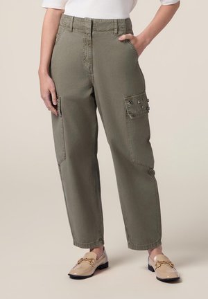 Personne portant un pantalon cargo vert olive avec des poches latérales à clous et des mocassins beige avec des détails en chaîne dorée.