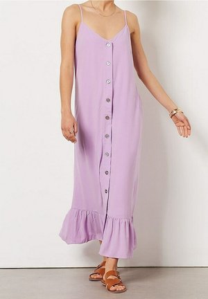 Kvinde iført en ærmeløs lilla midikjole med knapper foran og flæsekant samt brune flade sandaler, stående op ad en hvid væg.