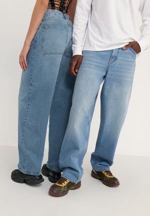 Ένα άτομο με φαρδιά μπλε τζιν και μαύρα αθλητικά παπούτσια, το άλλο με πιο ανοιχτόχρωμα φαρδιά μπλε τζιν, λευκό μακρυμάνικο πουκάμισο και πολύχρωμα αθλητικά παπούτσια.