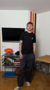 Jeune homme tatoué portant un t-shirt noir et un pantalon, debout dans une pièce avec une télévision, une lampe orange, un ballon de basket, des livres et un coffre de rangement.