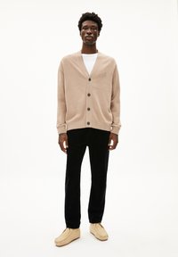 Beige cardigan met een V-hals, knopen aan de voorkant en ribgebreide mouwen, gecombineerd met zwarte broek en lichte schoenen. Glad gebreide textuur.