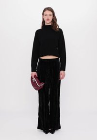Schwarzer, kurzer Pullover mit hohem Kragen, kombiniert mit schwarzen Velours-Hose in weitgeschnittenem Stil. Das Model hält eine kastanienbraune Clutch-Tasche mit silbernen Beschlägen.