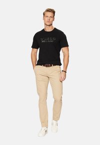 Camiseta de algodón negra con el logo de "GUESS", pantalones de algodón beige, cinturón marrón y zapatillas blancas. El modelo está de pie contra un fondo liso.