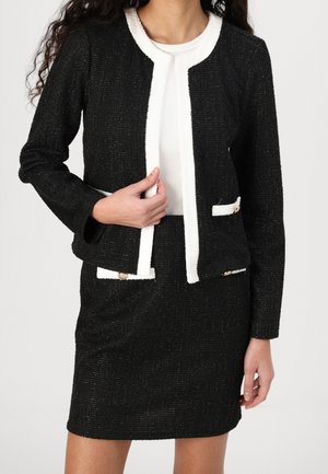 Femme vêtue d'un tailleur jupe noir texturé avec des finitions blanches et des boutons dorés, associé à un top blanc côtelé en dessous.