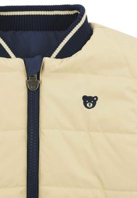 Giacca trapuntata beige con collo a coste blu navy, chiusura con zip e piccolo logo di un orsetto blu navy sul petto sinistro. Texture liscia con accenti a contrasto.