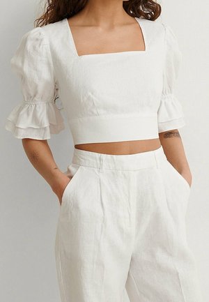 Woman wearing a white linen crop top with puffed sleeves and high-waisted white linen trousers, hands in pockets, tattoos visible on forearms.