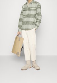 Camisa de cuadros verde de botones, pantalones cargo crema con bolsillos laterales, bolsa de tote beige con asas grises y botines beige.