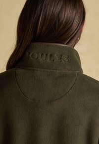 Person with long brown hair wearing an olive green fleece jacket with "COULES" embroidered on the collar, shown from the back.