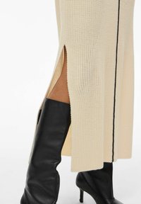 Robe midi crème en tricot avec fentes sur les côtés, détail de lignes verticales noires, associée à des bottes ajustées noires au-dessus du genou. Tissu texturé.