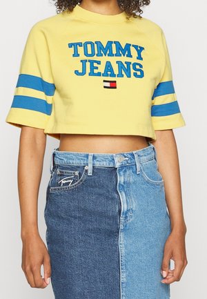 Persona que lleva un top corto amarillo con el texto "Tommy Jeans" y rayas azules en las mangas, combinado con una falda vaquera bicolor en tonos oscuros y claros.