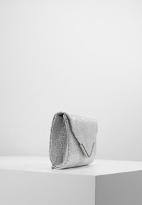 Sac à main argent métallisé avec une surface texturée, rabat repliable et bandoulière en chaîne. Présente un accent en matériel argenté minimaliste.