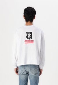 White long-sleeve t-shirt featuring a graphic print on the back, with a black and red design, paired with light blue jeans.