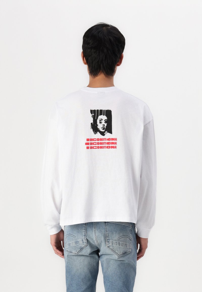 White long-sleeve t-shirt featuring a graphic print on the back, with a black and red design, paired with light blue jeans.