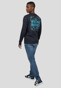 Svart tröja med en turkos grafisk textdesign som står "The WRONG FRIENDS" på baksidan. Bärs med blå jeans och svarta sneakers.