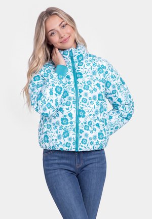 Junge Frau trägt eine weiße und türkisfarbene, blumige Reißverschlussjacke, steht mit einer Hand am Hals und der anderen in der Tasche.