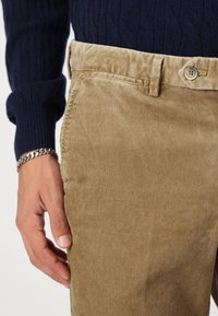 Pantalon en velours côtelé couleur beige avec une finition texturée, présentant une fermeture à bouton et des coutures le long de la taille, porté avec un pull bleu marine.