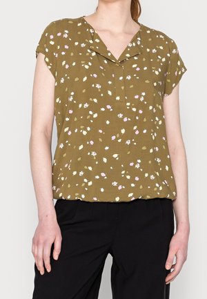 Blouse vert olive à manches courtes avec un petit motif floral blanc et rose, portée avec un pantalon noir, vue mi-corps sur fond clair.