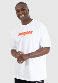 Hvid bomulds T-shirt med korte ærmer og rund halsudskæring, med et orange "SMILODOX" logo på fronten. Sort tekstdetalje i bunden.