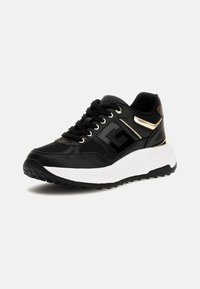 Basket de sport noire et dorée avec une épaisse semelle blanche, des lacets noirs, une tige en maille et cuir, et un talon texturé surélevé, inclinée vers la droite sur fond blanc.