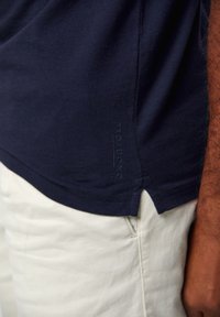 Navyblaue Baumwoll-T-Shirt mit seitlichem Schlitz, versehen mit eingeprägtem Branding am Saum. Kombiniert mit hellen, strukturierten Hosen.