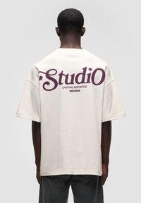 Creme oversized bomulds T-shirt med stor purple tekst på ryggen: "Studio" og "CRAFTING AESTHETICS WRSTBHVR." Korte ærmer.