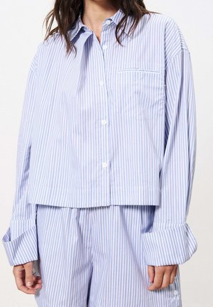 Femme portant une chemise ample à rayures verticales bleu clair et blanc avec boutons, un pantalon ample assorti et les manches retroussées.