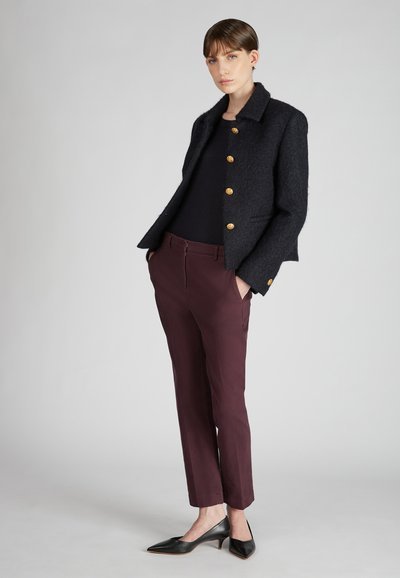 Veste en tissu texturé bleu marine avec boutons dorés, associée à un pantalon ajusté bordeaux et des escarpins noirs à bout pointu, créant un look sur mesure.