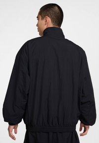 Svart jakke med høy krage, elastiske mansjetter og midjebånd. Laget av lett materiale med løs passform og glatt tekstur.