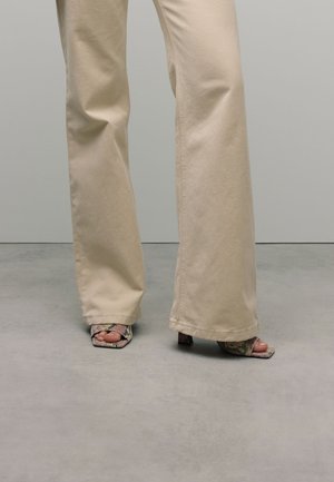 Beige weite Hose mit glatter Textur, kombiniert mit offenen Schuhen mit Schlangenleder-Muster und einem kleinen Blockabsatz.