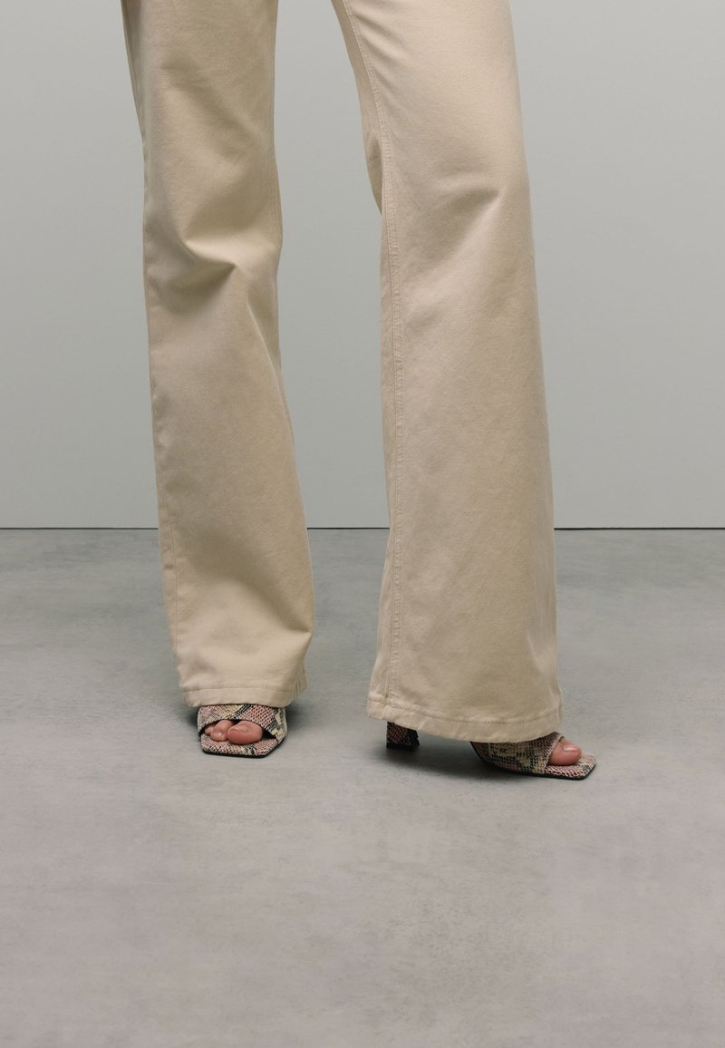 Pantalons larges beiges à texture lisse, associés à des chaussures ouvertes au bout avec un motif serpent et un petit talon carré.