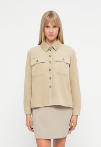 Μπεζ πουκάμισο ζακέτα από κοτλέ με δύο τσέπες στο στήθος, κλείσιμο με κουμπιά και κολάρο, συνδυασμένο με μια ταιριαστή φούστα.