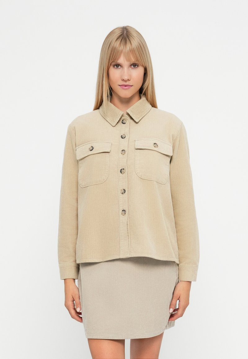 Μπεζ πουκάμισο ζακέτα από κοτλέ με δύο τσέπες στο στήθος, κλείσιμο με κουμπιά και κολάρο, συνδυασμένο με μια ταιριαστή φούστα.